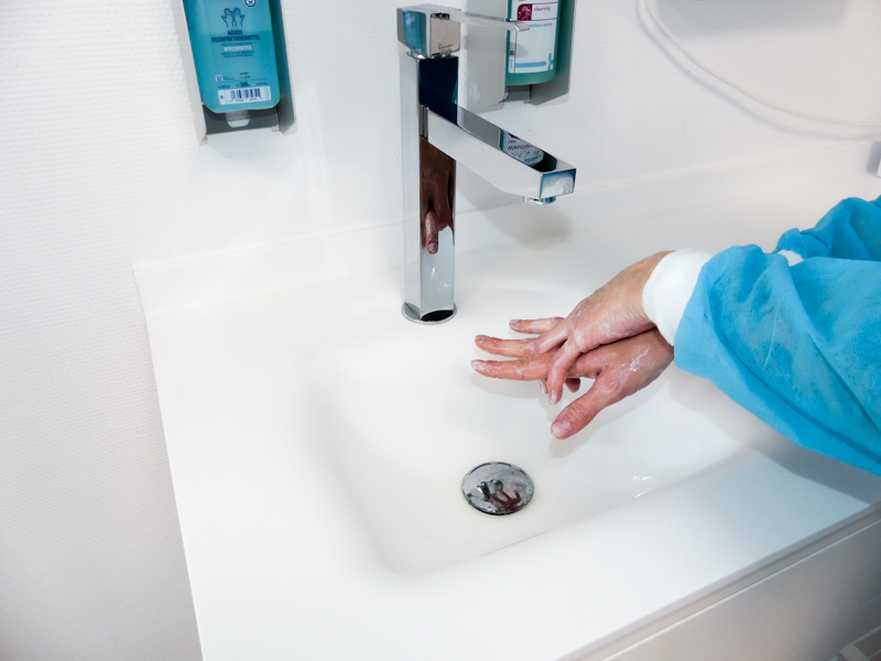 Person in blauer Schutzkleidung wäscht Hände unter einem Wasserhahn an einem weißen Waschbecken.