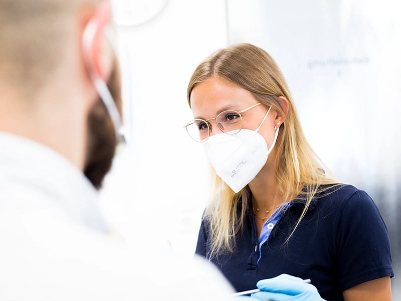 Eine Ärztin mit FFP2-Maske und Brille führt eine Untersuchung bei einem Patienten mit Maske durch.