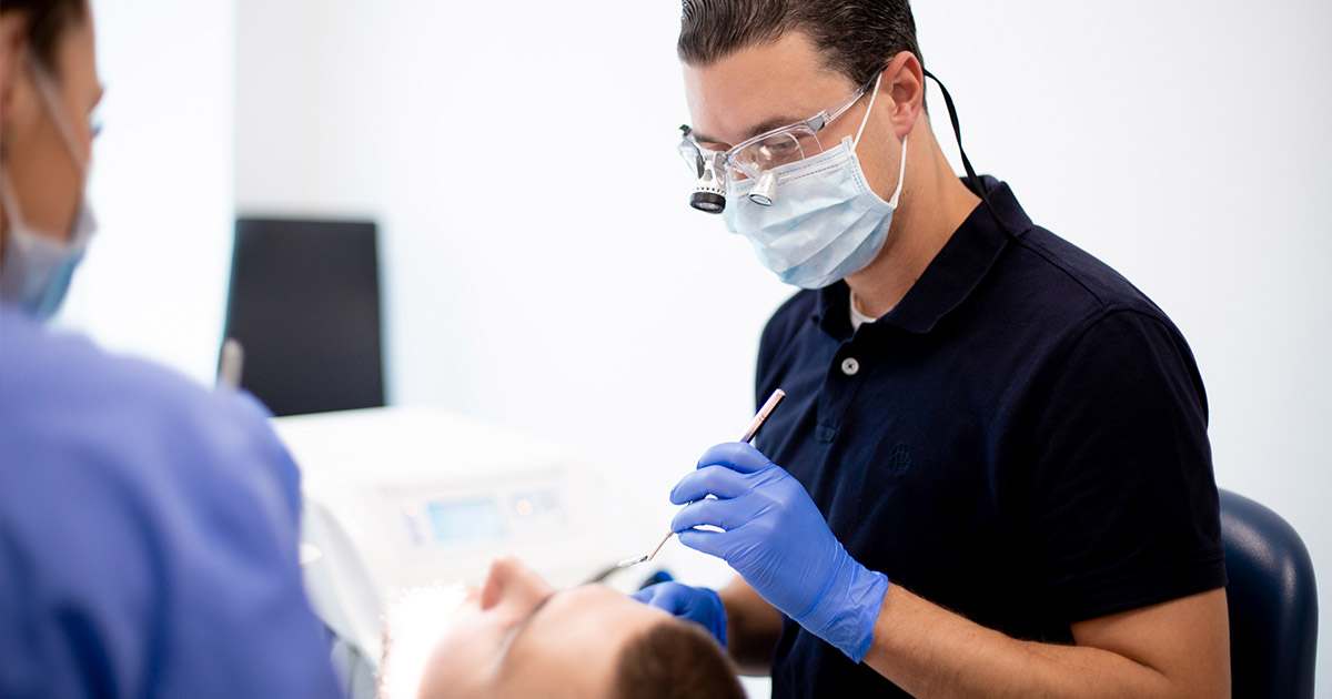 Zahnarzt in dunkler Kleidung untersucht einen Patienten im Behandlungsstuhl, trägt Mundschutz, Schutzbrille und Handschuhe.